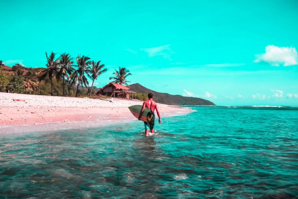 The back of a surfer walking through the water with his board on to Kuta beach