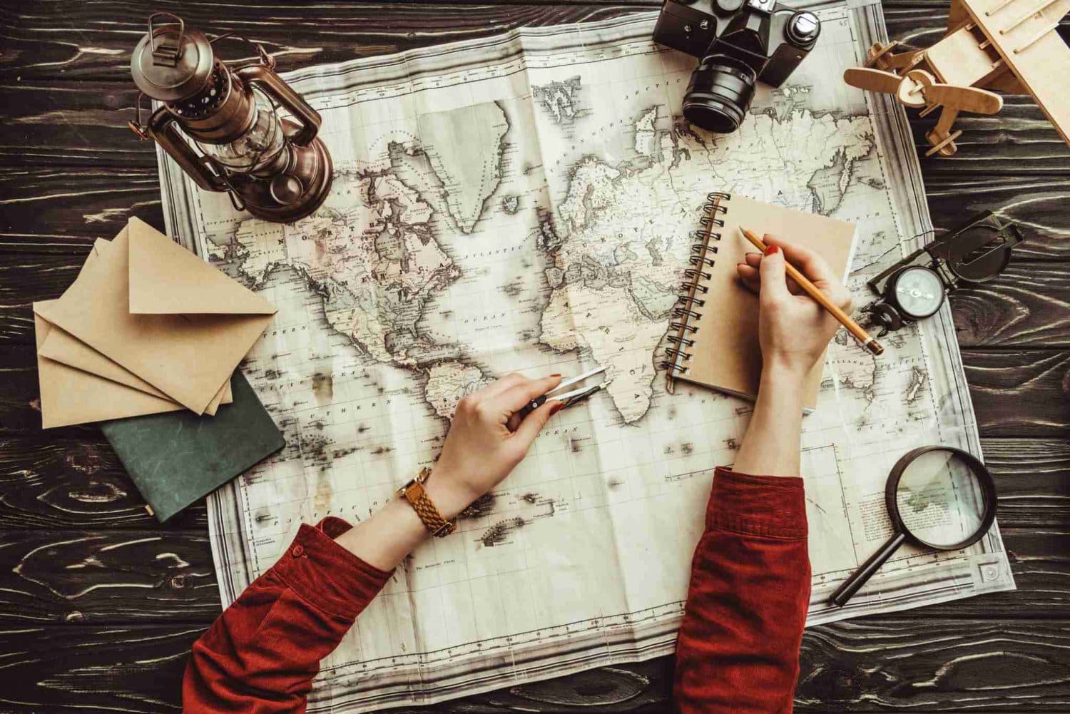 Birds' eye view of a woman in a red sweater recording her travel memories with a journal, map and postcards