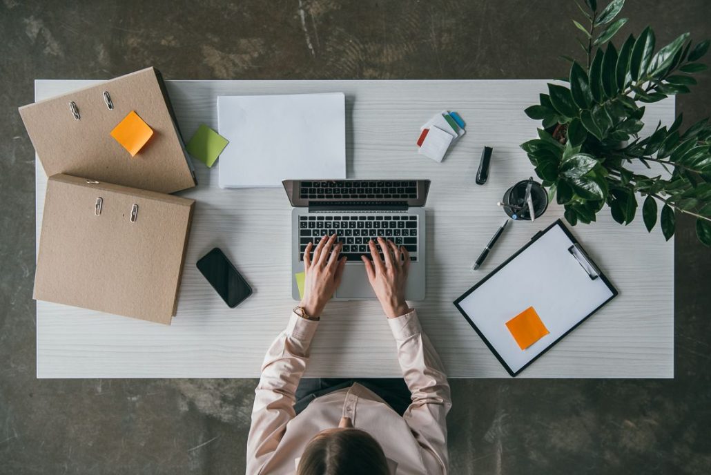 Top view photo of a woman working at a desk with a laptop, some binders, her cellphone, a clipboard and plant.