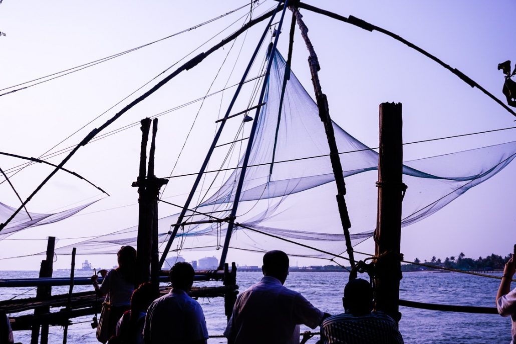 Fishers surrounding their set-up Chinese fishing nets. The nets are strung along a frame created by four posts that are connected at the top in a tent shape.