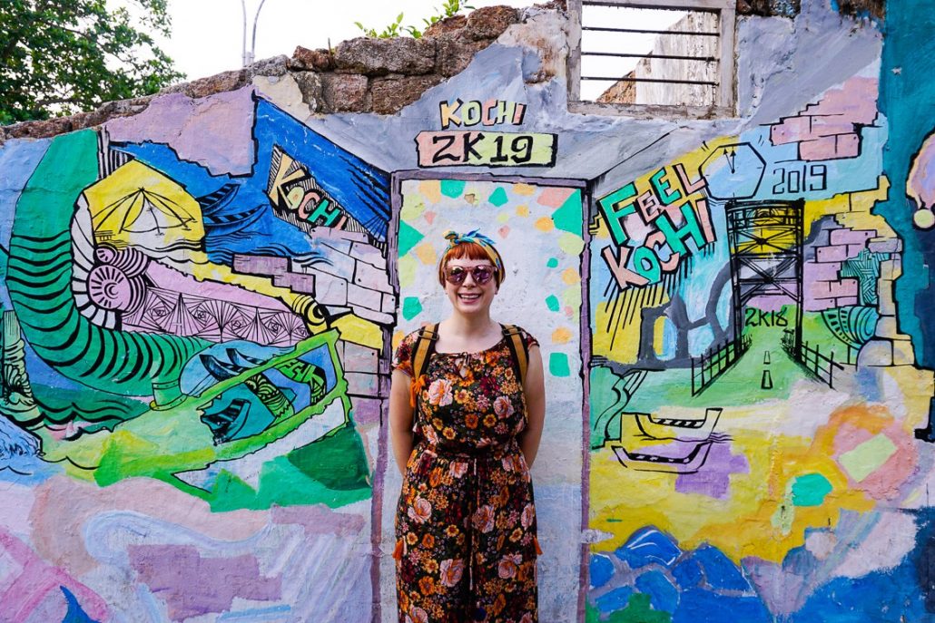 Alice smiling and standing against a ruined building in Fort Kochi covered in blue, yellow and green graffiti.