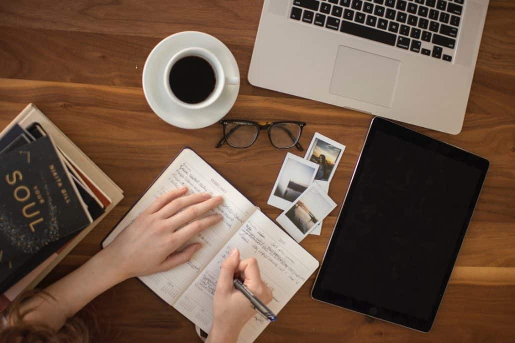 Top down photo of a person writing in a planner with an iPad, Macbook, photos, glasses, coffee, and books around them.