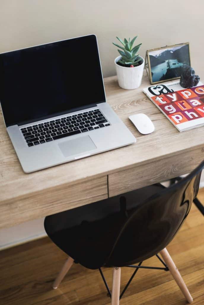 Photo of a laptop in a desk with a black chair tucked underneath. Some other decorations include a plant, some books, and a photo frame.