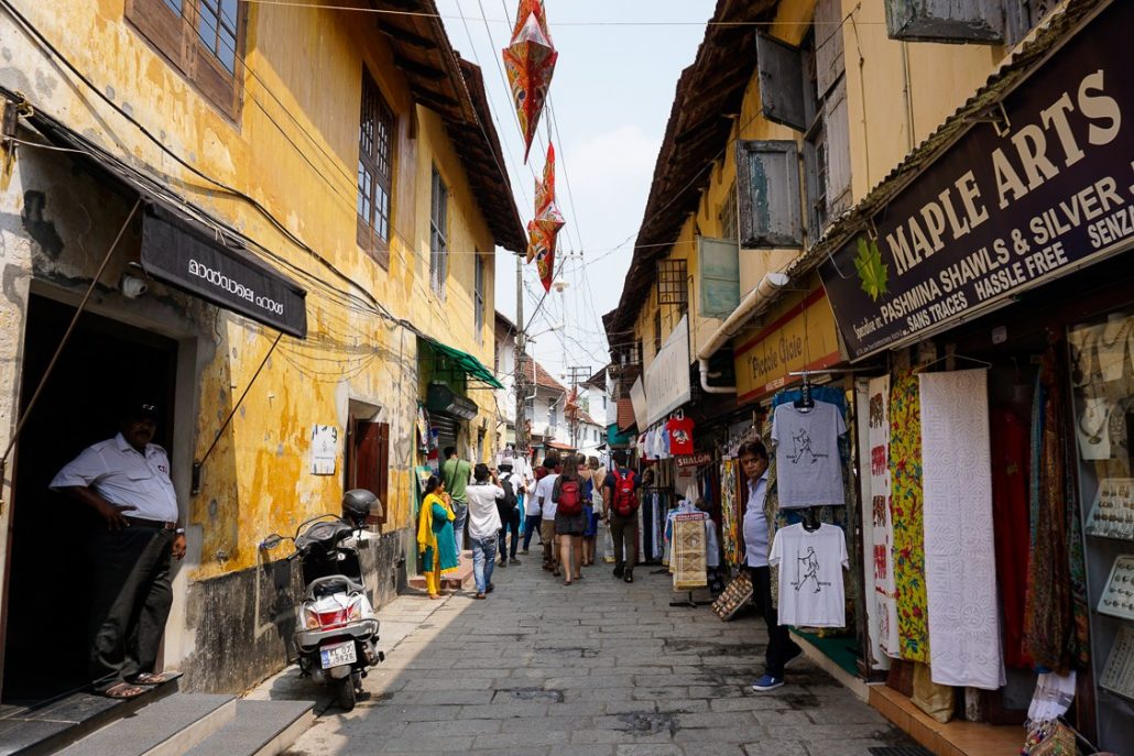 A busy, narrow street with shopkeepers on each side standing outside their shops. They sell pashminas and other clothing items.