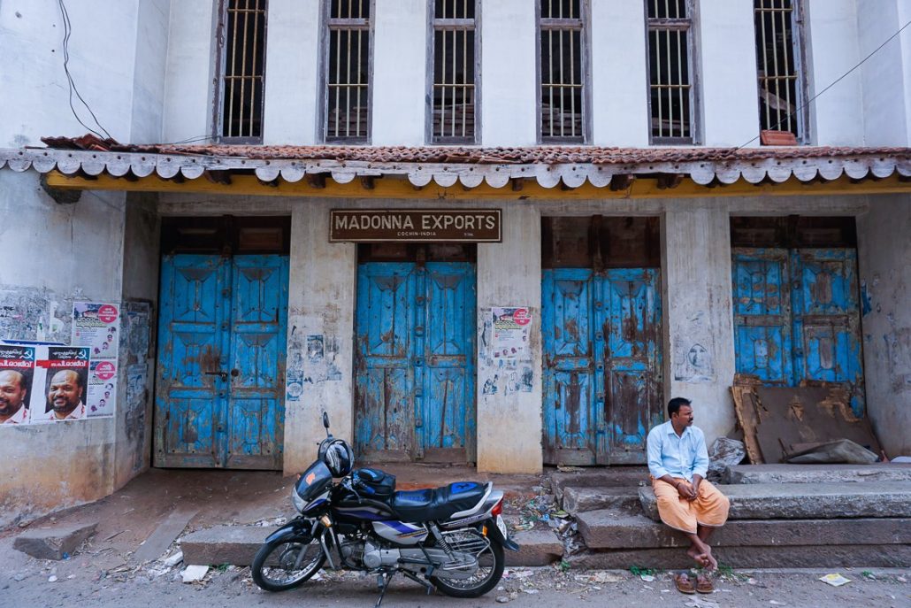 An old, white building featuring four blue double doors and a sign that says "MADONNA EXPORTS". There is also a motorcycle in the middle of the frame with a man resting to the right of it.