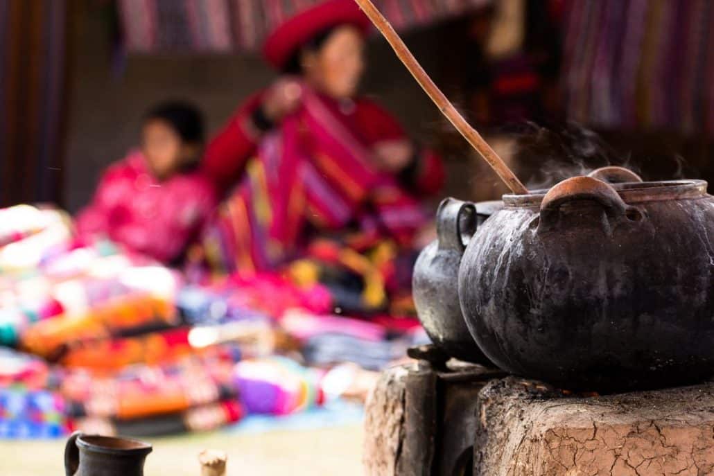 Photo with two steaming pots in the foreground with out of focus woman and daughter behind a bunch of brightly coloured fabrics.