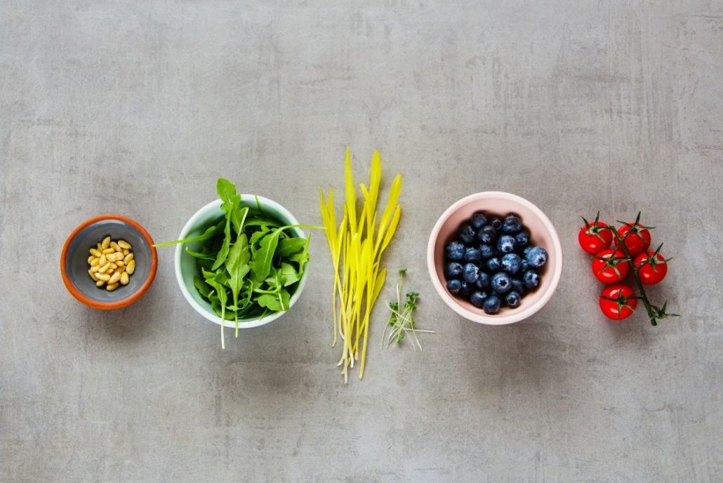 Various raw organic food arugula, cress salad, blueberries, cherry tomatoes, pine nuts over light background